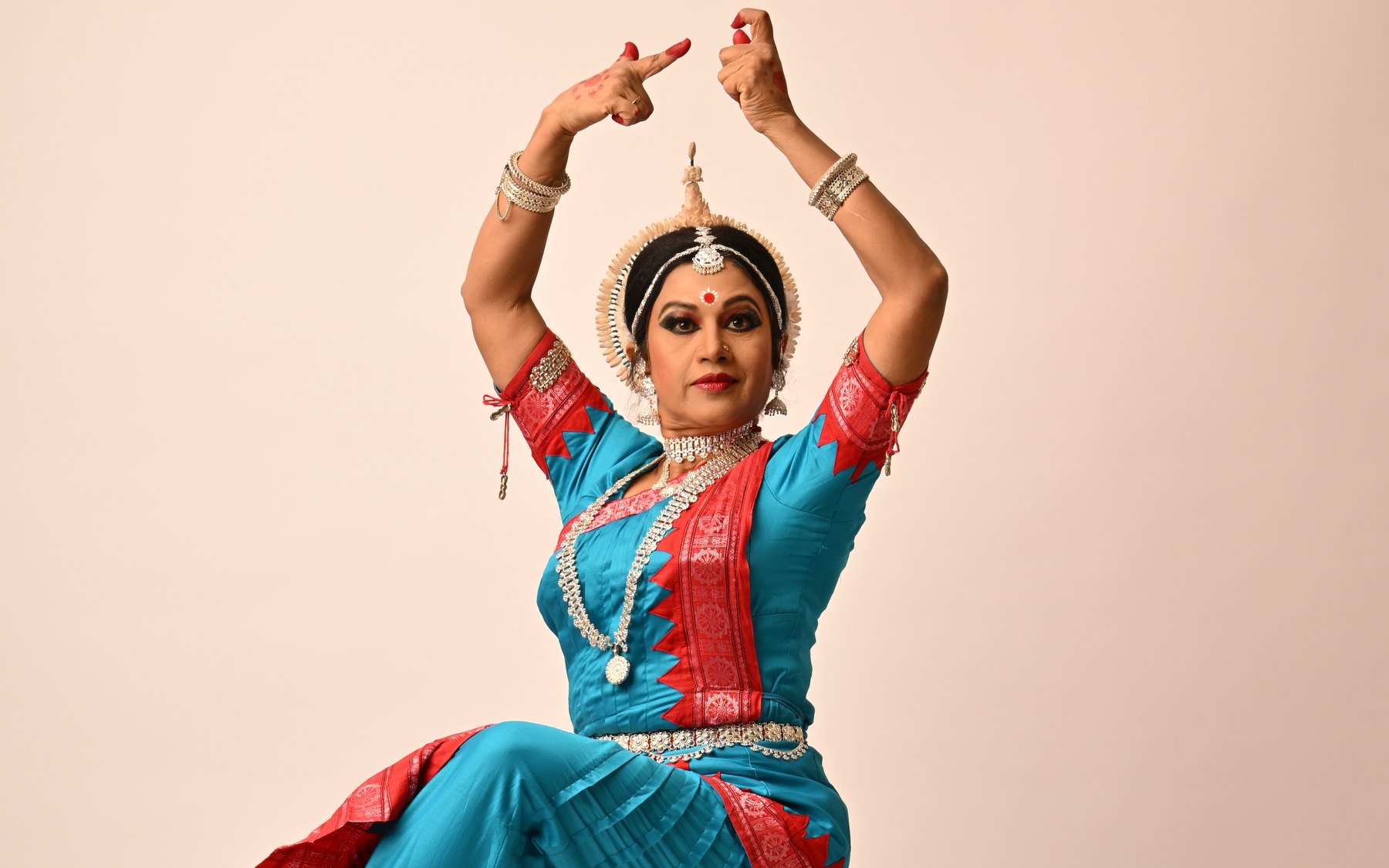 Shubhada Varadkar in an Odissi pose, arms raised above her head, in a teal and red costume.