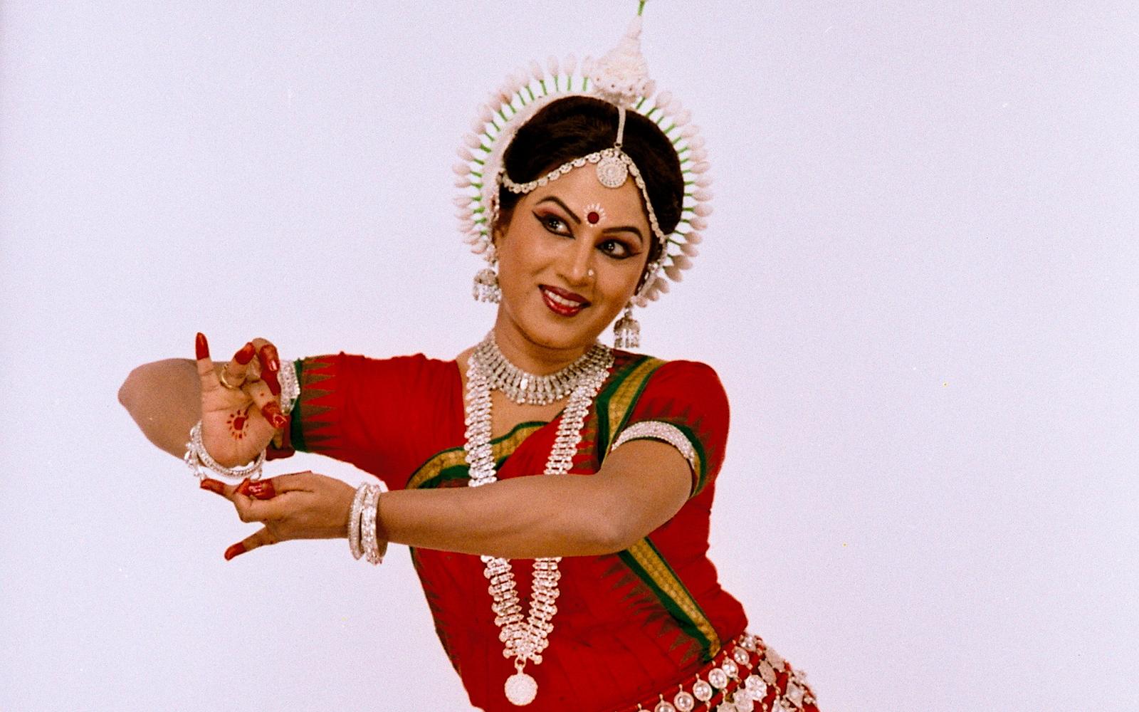 Shubhada Varadkar in a red Odissi costume, arms extended to the side.
