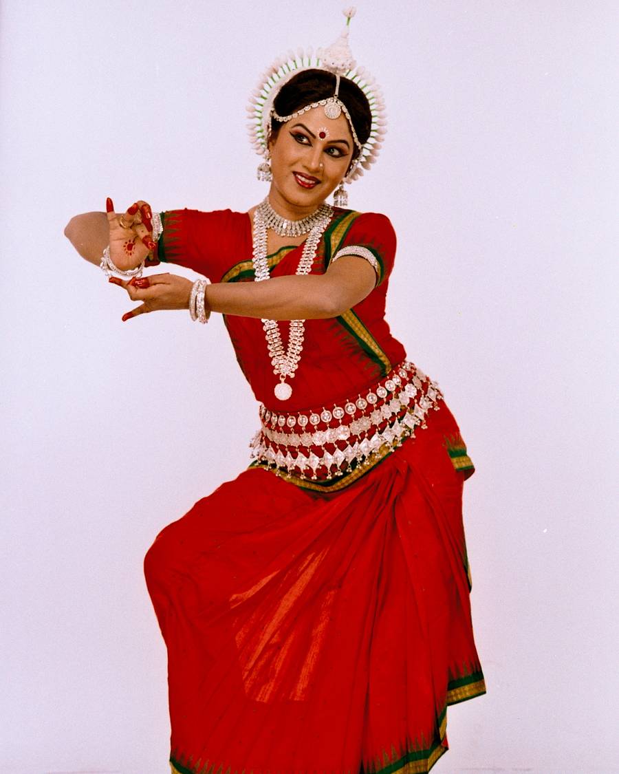 Shubhada Varadkar in a red Odissi costume, arms extended to the side.