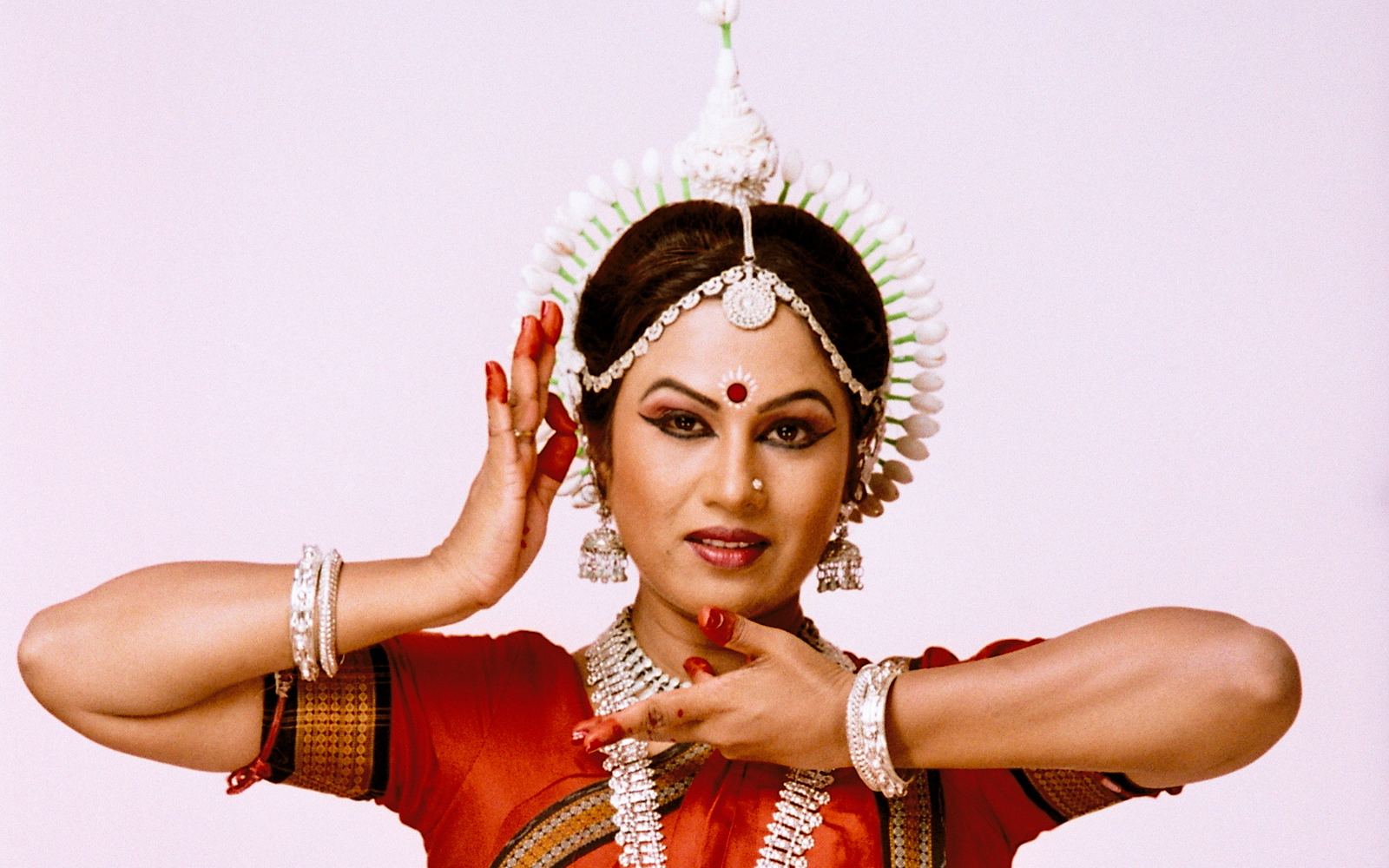 Shubhada Varadkar in a red Odissi costume, hands raised beside her face.