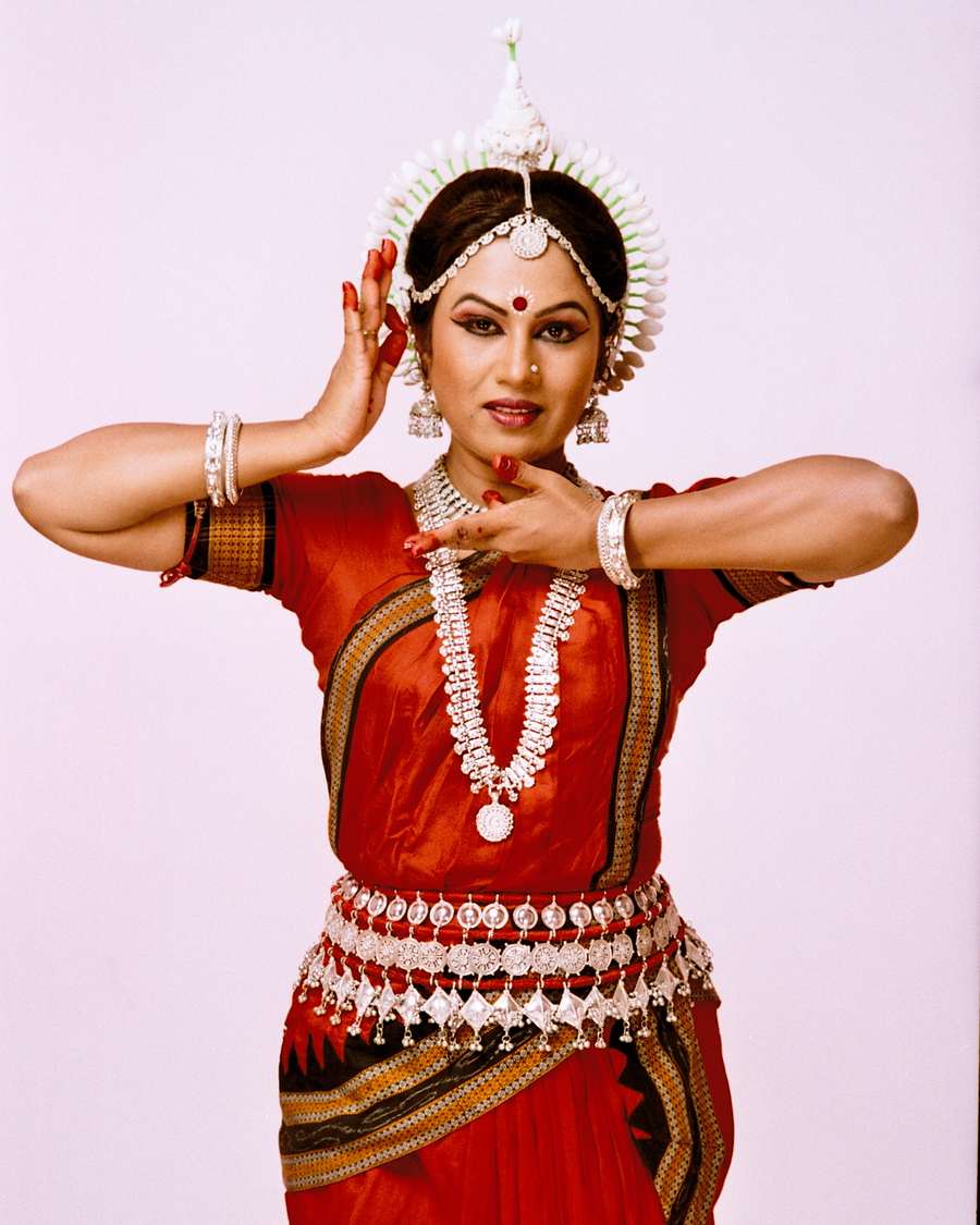 Shubhada Varadkar in a red Odissi costume, hands raised beside her face.