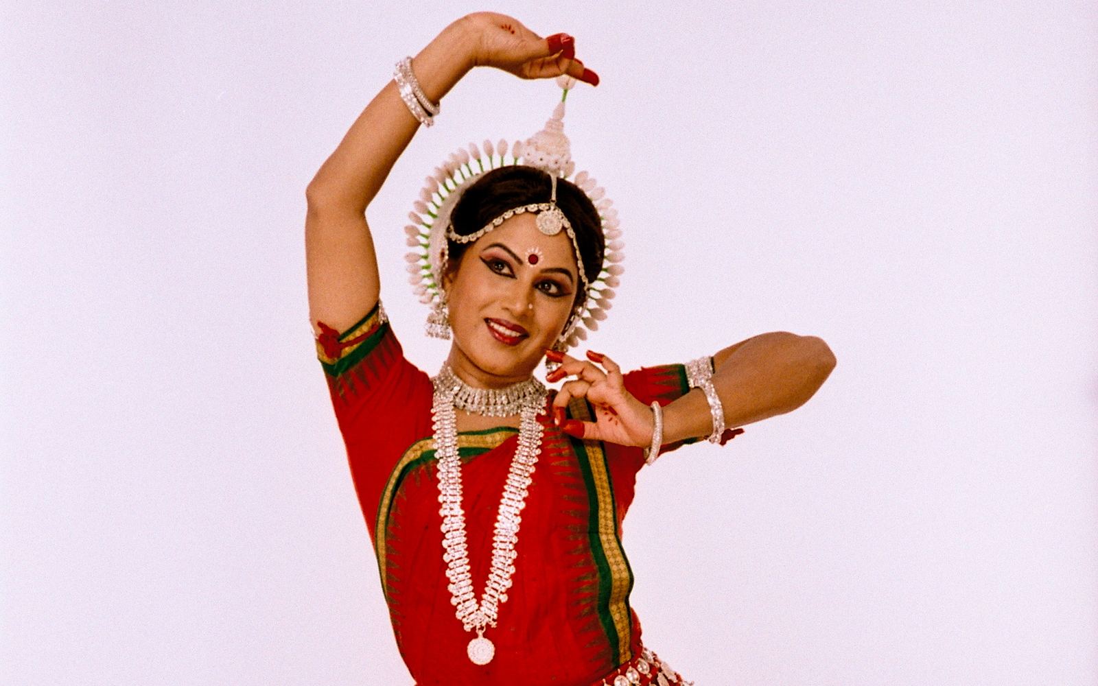 Shubhada Varadkar in a red Odissi costume, one arm lifted overhead.