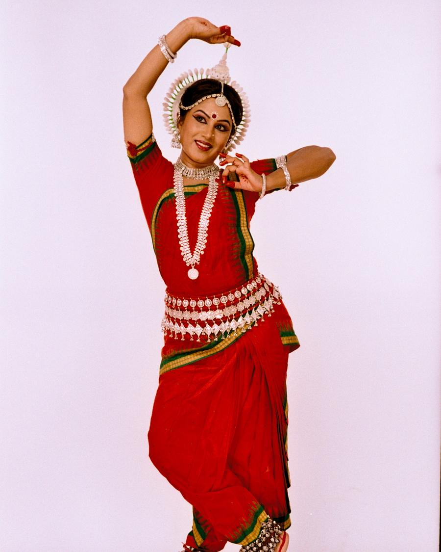 Shubhada Varadkar in a red Odissi costume, one arm lifted overhead.