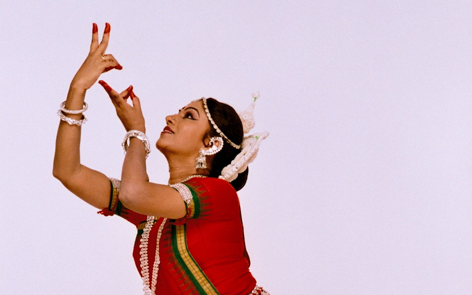 Shubhada Varadkar in a red Odissi costume, one leg lifted mid-movement.