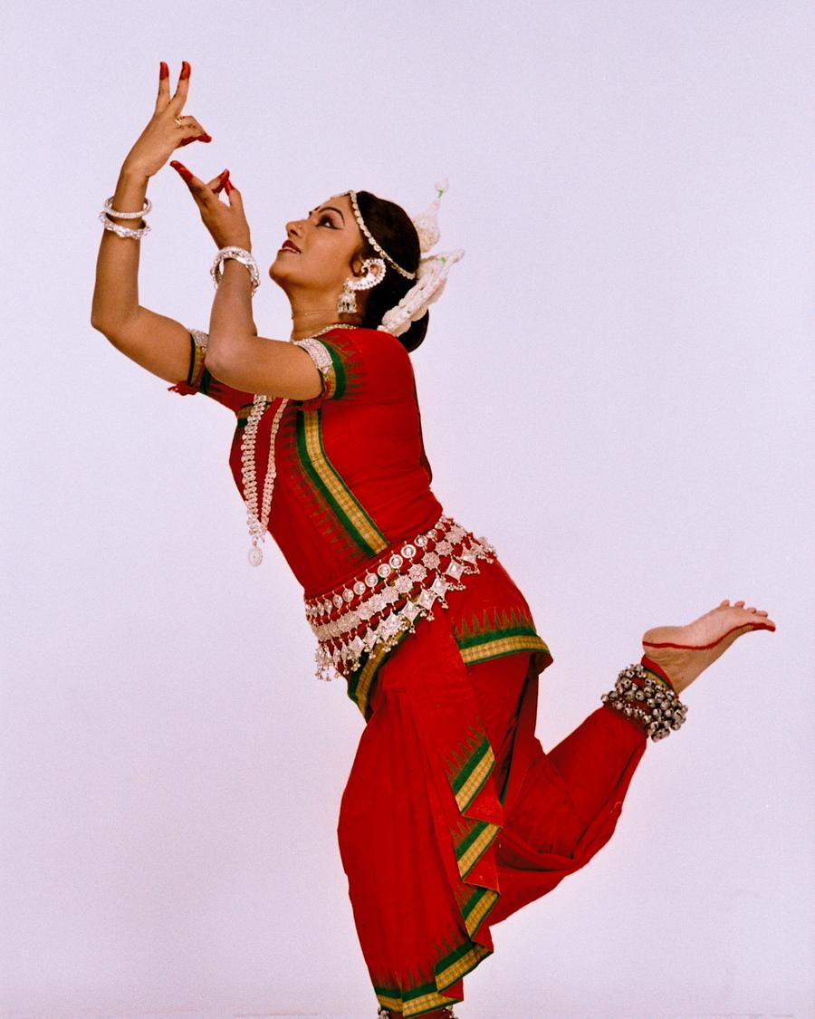 Shubhada Varadkar in a red Odissi costume, one leg lifted mid-movement.